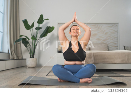 Yoga mindfulness meditation. Woman practicing yoga in living room at home. Plus size female sitting in lotus pose meditating smiling relaxing indoor. Girl doing breathing practice. Yoga at home 132834565