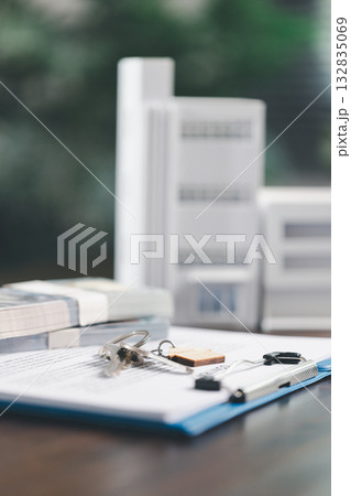 Real estate agent and customer are discussing contract to buy house, with house model in background symbolizing real estate, insurance, and loan arrangements involved in process. 132835069