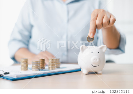 Concept of saving money and investment planning. A businesswoman drops coin into white piggy bank on office desk with documents, symbolizing financial security, budget management and future wealth. 132835112