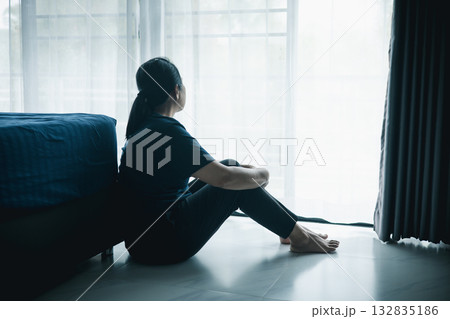 A lonely woman sits on the floor by window looking outside in dark room. Concept of depression, anxiety, sadness, loneliness, mental health problem, emotional struggle and hope for better life. A lonely woman sits on the floor by window looking outside in dark room. Concept of depression, anxiety, sadness, loneliness, mental health problem, emotional struggle and hope for better life. 132835186