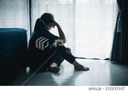 A lonely woman sits on the floor by window with hand on head in dark room. Concept of depression, sadness, anxiety, stress, mental health problem, loneliness and emotional struggle in life. A lonely woman sits on the floor by window with hand on head in dark room. Concept of depression, sadness, anxiety, stress, mental health problem, loneliness and emotional struggle in life. 132835187