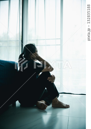 A lonely woman sits on the floor by window with hand on head in dark room. Concept of depression, sadness, anxiety, stress, mental health problem, loneliness and emotional struggle in life. A lonely woman sits on the floor by window with hand on head in dark room. Concept of depression, sadness, anxiety, stress, mental health problem, loneliness and emotional struggle in life. 132835188