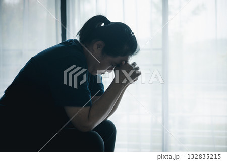 A stressed woman sitting alone on bed in dark room, holding her head down, symbolizing depression, anxiety, hopelessness, loneliness, mental health struggle, burnout and emotional pain. 132835215