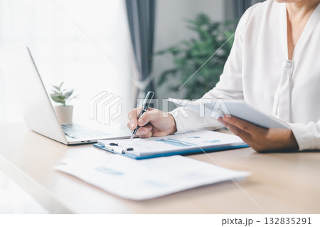 A businesswoman writes notes on clipboard while holding notebook at office desk with documents. Concept of business planning, accounting, financial management, productivity and corporate strategy. 132835291