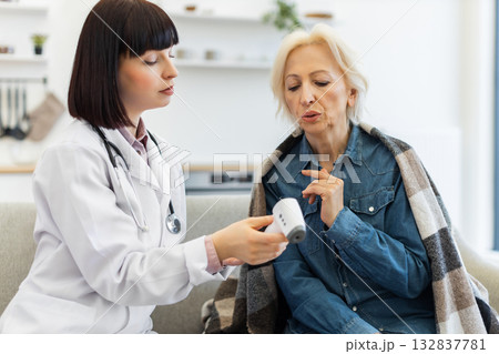 A female doctor checks the temperature of an elderly woman using a digital thermometer during a home visit. The patient is wrapped in a blanket. A female doctor checks the temperature of an elderly woman using a digital thermometer during a home visit. The patient is wrapped in a blanket. 132837781