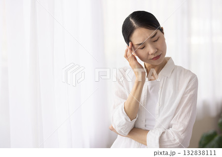 窓辺でこめかみに手を当て眉間に皺を寄せるミドル女性 窓辺でこめかみに手を当て眉間に皺を寄せるミドル女性 132838111