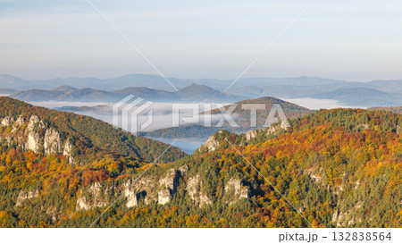 Vibrant autumn foliage blankets mountainous terrain, as a layer of mist softens the distant peaks under a serene sky. The Sulov Rocks, national nature reserve in northwest of Slovakia, Europe. Vibrant autumn foliage blankets mountainous terrain, as a layer of mist softens the distant peaks under a serene sky. The Sulov Rocks, national nature reserve in northwest of Slovakia, Europe. 132838564