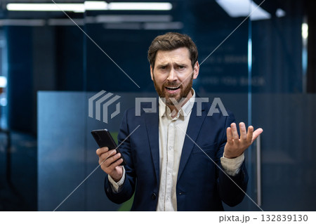 Portrait of a worried young businessman in a suit standing in the office, holding a phone and spreading his arms in frustration at the camera. 132839130