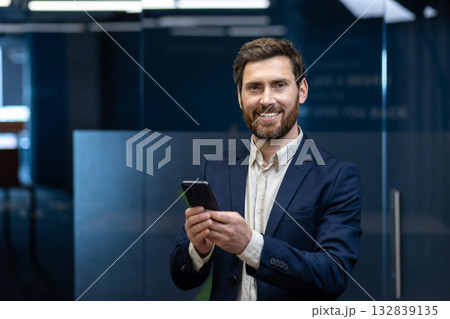 Portrait of a smiling young businessman in a suit standing in an office, holding a phone, looking at the camera. 132839135