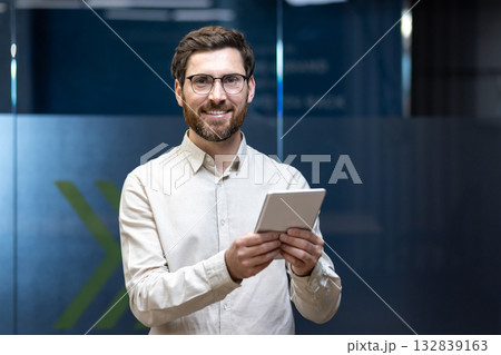 Close-up portrait of a young smiling man standing in the office with a tablet in his hands and looking at the camera. 132839163