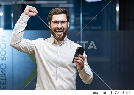 Portrait of a happy young man in a company office, holding a phone and celebrating success with his hands raised. Portrait of a happy young man in a company office, holding a phone and celebrating success with his hands raised. 132839168