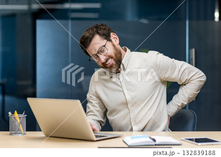Tired young male office worker sitting at desk, holding hand behind back and suffering from lower back pain. Tired young male office worker sitting at desk, holding hand behind back and suffering from lower back pain. 132839188