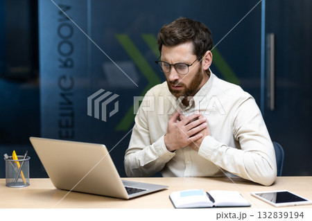 A young businessman grimacing in pain sits at a desk in the office and holds his hands to his chest. 132839194
