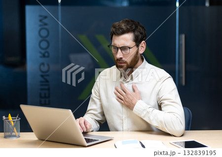 A young male businessman sits at a desk in the office and holds his hand to his chest, feeling strong pressure and pain. A young male businessman sits at a desk in the office and holds his hand to his chest, feeling strong pressure and pain. 132839195