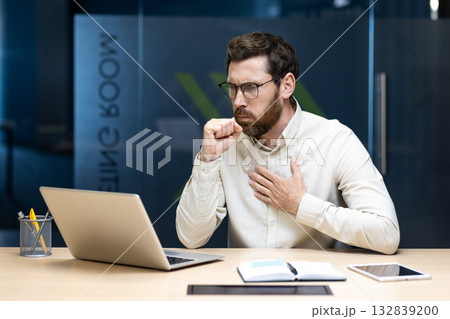 A young male office worker sits at a desk in the office, holds his hand to his chest and experiences severe coughing fits. 132839200