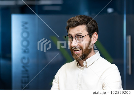 Close-up portrait of a smiling young man in glasses and shirt standing in the office, looking and smiling at the camera. Close-up portrait of a smiling young man in glasses and shirt standing in the office, looking and smiling at the camera. 132839206