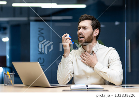A young businessman is puffing an inhaler down his throat, sitting at his desk in the office, and suffering from asthma. A young businessman is puffing an inhaler down his throat, sitting at his desk in the office, and suffering from asthma. 132839220