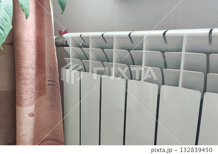 White modern radiator near a curtain and wall. Concept of home heating, comfort, and warmth during cold winter days. Cozy interior and energy efficiency in household design. White modern radiator near a curtain and wall. Concept of home heating, comfort, and warmth during cold winter days. Cozy interior and energy efficiency in household design. 132839450