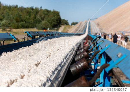 Long blue conveyor belt transports white potash ore material toward massive potassium mining dump sunlight. Industrial machinery stretches distance mineral extraction continuous production process 132840957