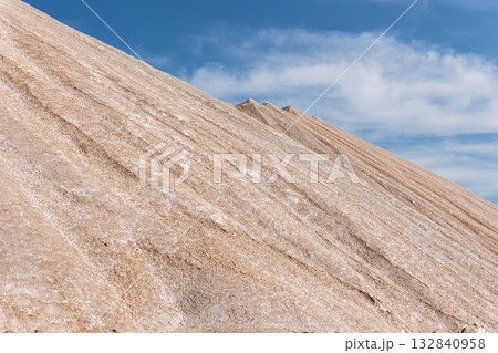 Potassium ore dump. Massive mound potash waste rises deep blue sky, its surface marked by mineral lines erosion patterns. Arid landscape sparse vegetation industrial origin and stark natural contrast 132840958