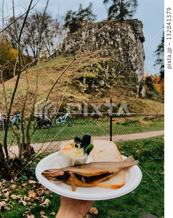 Hand holding disposable plate featuring smoked trout, bread, salad with Sokolica peak backdrop in Ojcowski National Park during colorful autumn landscape. Hand holding plate with smoked trout Hand holding disposable plate featuring smoked trout, bread, salad with Sokolica peak backdrop in Ojcowski National Park during colorful autumn landscape. Hand holding plate with smoked trout 132841379