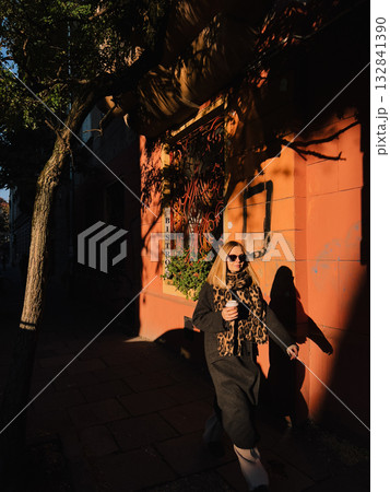 Fashionable blonde strolling bistro sidewalk during golden hour, drinking takeaway coffee, wearing long dark coat and leopard print scarf, radiating sophisticated urban style. Stylish blonde woman 132841390