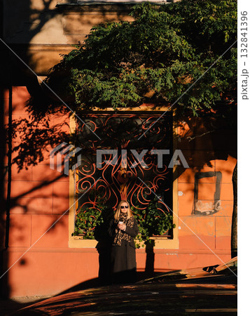 Blonde woman in sunglasses and coat, holding coffee cup, stands near old building with decorative window, bathed in warm glow of afternoon sun. Portrait of blonde woman wearing sunglasses and coat 132841396