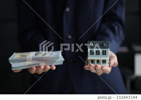 Businessman holding a stack of money in one hand and a miniature house in the other. real estate investment concept. purchasing, owning, managing, renting, or selling real estate to generate profit or 132843194