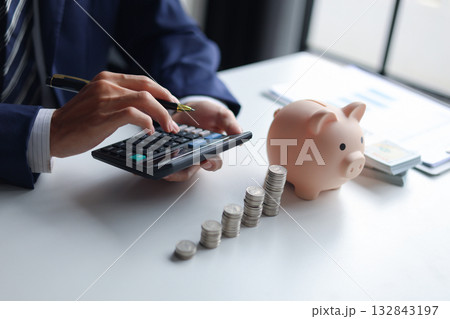 Businessman using a calculator with a piggy bank, stacks of coins, and US dollar bills on a table. Concept of saving money and financial planning. Businessman using a calculator with a piggy bank, stacks of coins, and US dollar bills on a table. Concept of saving money and financial planning. 132843197