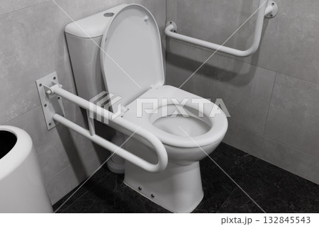 A toilet in a public or accessible restroom featuring safety grab bars (rails) mounted on the gray tiled wall. A toilet in a public or accessible restroom featuring safety grab bars (rails) mounted on the gray tiled wall. 132845543