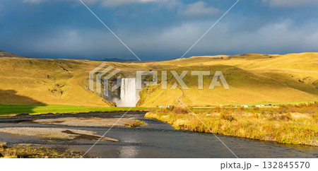 Skogafoss waterfall in Iceland cascades down a steep cliff, creating a powerful mist that forms a vibrant rainbow. The surrounding lush green landscape enhances its majestic beauty Skogafoss waterfall in Iceland cascades down a steep cliff, creating a powerful mist that forms a vibrant rainbow. The surrounding lush green landscape enhances its majestic beauty 132845570