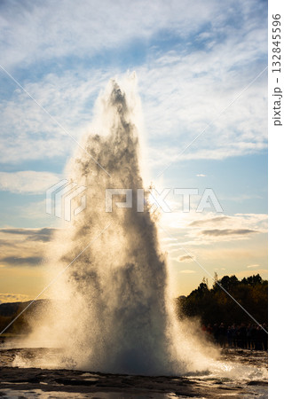 Strokkur geyser erupts in Iceland, sending a plume of water and steam high into the air. Tourists gather nearby, capturing the spectacle against a dramatic sky. Strokkur geyser erupts in Iceland, sending a plume of water and steam high into the air. Tourists gather nearby, capturing the spectacle against a dramatic sky. 132845596