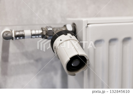 A close-up of a white thermostatic radiator valve (TRV) and its control knob on a modern panel radiator against a gray tiled wall. 132845610