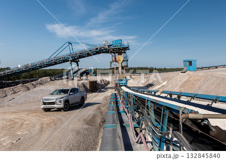 Bright sunlight illuminates open industrial potassium mining site blue conveyor structures silver pickup truck car dusty ground. Steel constructions contrast with light rocks and clear blue sky Bright sunlight illuminates open industrial potassium mining site blue conveyor structures silver pickup truck car dusty ground. Steel constructions contrast with light rocks and clear blue sky 132845840