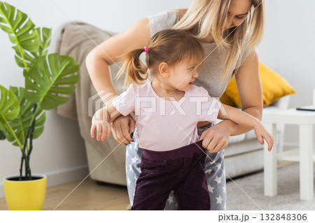 Mother dressing her playful daughter at home on a sunny morning. Joyful parenting, affection, and the simple happiness of family routine. 132848306