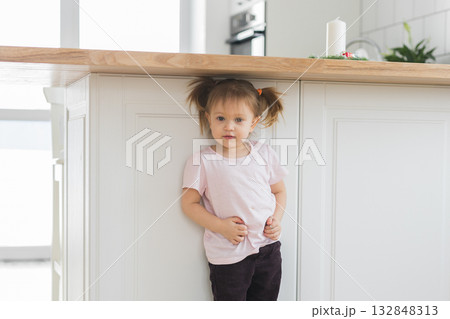 Little girl standing by the kitchen counter in soft daylight. Innocence, curiosity, and the calm rhythm of childhood in a modern home interior. 132848313