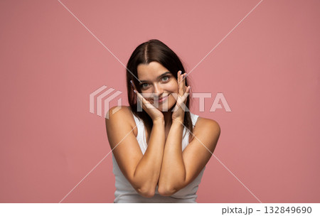 Playful young woman posing with hands on cheeks smiling gently against pastel pink background 132849690