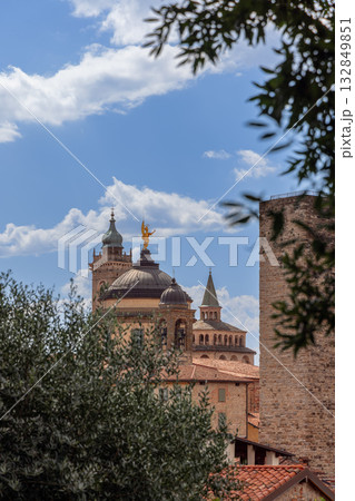Bergamo skyline of domes towers and Saint Alexander statue glowing above roofs and green leaves 132849851