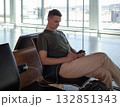 Young man charging phone at airport gate, using smartphone while waiting for flight. Comfortable travel concept in modern terminal. 132851343