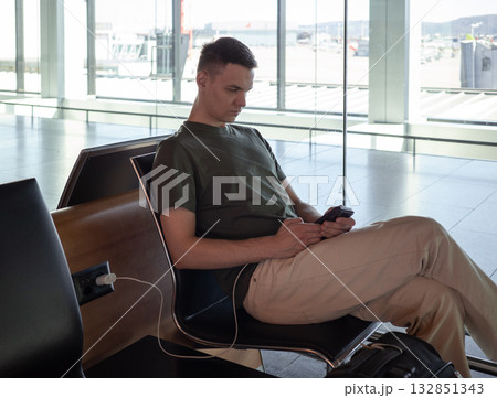 Young man charging phone at airport gate, using smartphone while waiting for flight. Comfortable travel concept in modern terminal. Young man charging phone at airport gate, using smartphone while waiting for flight. Comfortable travel concept in modern terminal. 132851343