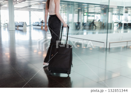 Young woman walking through airport terminal with black suitcase, motion blur. Concept of independent travel, modern lifestyle and seamless mobility. Photo in motion, soft focus. 132851344