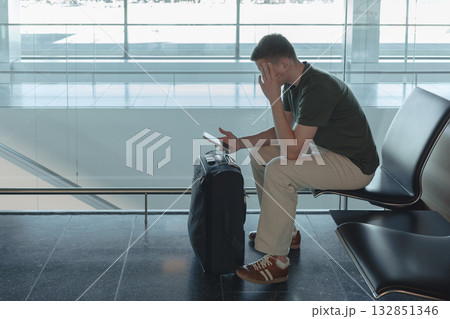 Young man sitting at airport gate with suitcase waiting for a flight, looking at his smartphone. Passenger is upset with flight delay. Modern air travel concept. Young man sitting at airport gate with suitcase waiting for a flight, looking at his smartphone. Passenger is upset with flight delay. Modern air travel concept. 132851346