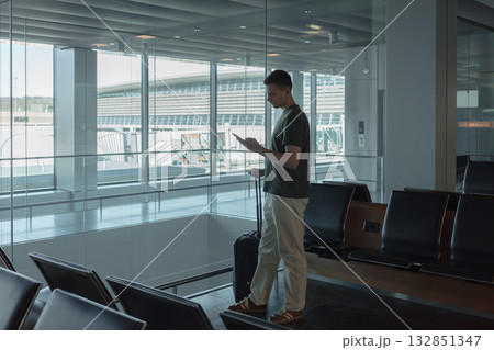 Man stands at a large airport window, holding his suitcase and checking his phone while waiting for his flight. Concept of solitude, anticipation and modern travel technology. 132851347