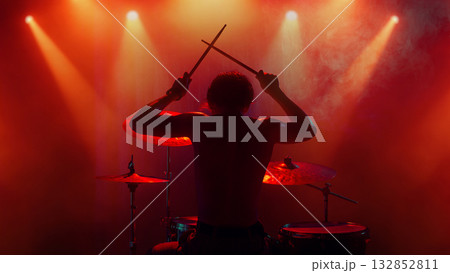 Drummer silhouette under orange light raising drumsticks in emotional finale Drummer silhouette under orange light raising drumsticks in emotional finale 132852811