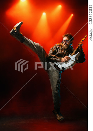 Dynamic musician kicking high while holding guitar under vivid red concert lights 132852833