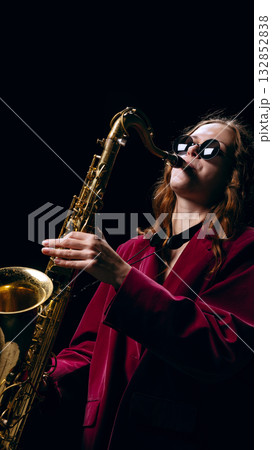 Woman playing golden saxophone wearing sunglasses and velvet jacket under stage light 132852838