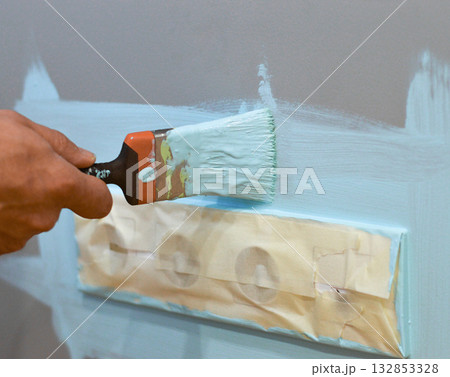 Applying paint to the wall surrounding an electrical socket using a brush, with masking tape for protection, as part of a DIY home improvement project, ensuring clean edges and safe interior upkeep. 132853328