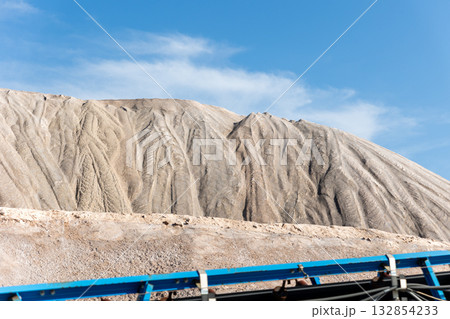 Long blue conveyor belt transports white potash ore material toward massive potassium mining dump sunlight. Industrial machinery stretches distance mineral extraction continuous production process Long blue conveyor belt transports white potash ore material toward massive potassium mining dump sunlight. Industrial machinery stretches distance mineral extraction continuous production process 132854233