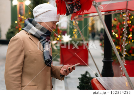 Mans buying red gift using smartphone at Christmas fairy. Xmas shopping. Side view. Holiday spirit. Close up. Mans buying red gift using smartphone at Christmas fairy. Xmas shopping. Side view. Holiday spirit. Close up. 132854253