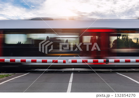 Fast modern regional commute train rushes past closed railway crossing under cloudy sky  sunlight sunset evening. Motion blur rush hour speed dynamic atmosphere  urban transportation commute scene 132854270